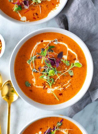 A close-up of a bowl of roasted red pepper soup topped with heavy cream and microgreens.