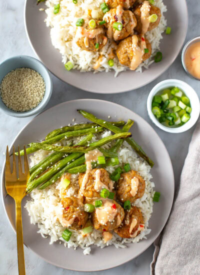 Overhead shot of two plates with firecracker meatballs, white rice, and green beans, garnished with green onions and sesame seeds. A gold fork rests on one plate.