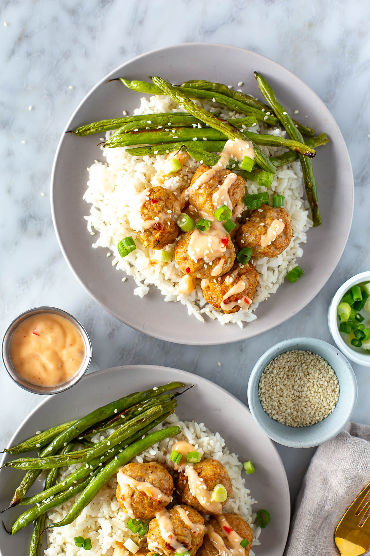 Overhead shot of two plates of firecracker meatballs with rice and green beans. Small bowls of sauce, sesame seeds, and chopped green onions are also visible on the marble surface.