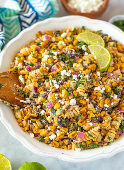 Overhead shot of Mexican street corn pasta salad in a white serving bowl with lime wedges and cilantro.