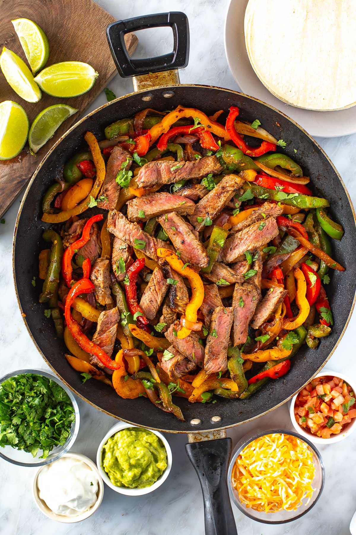 Overhead shot of steak fajitas in a skillet with bell peppers and onions, surrounded by bowls of guacamole, salsa, sour cream, shredded cheese, chopped cilantro, lime wedges, and a plate of tortillas.