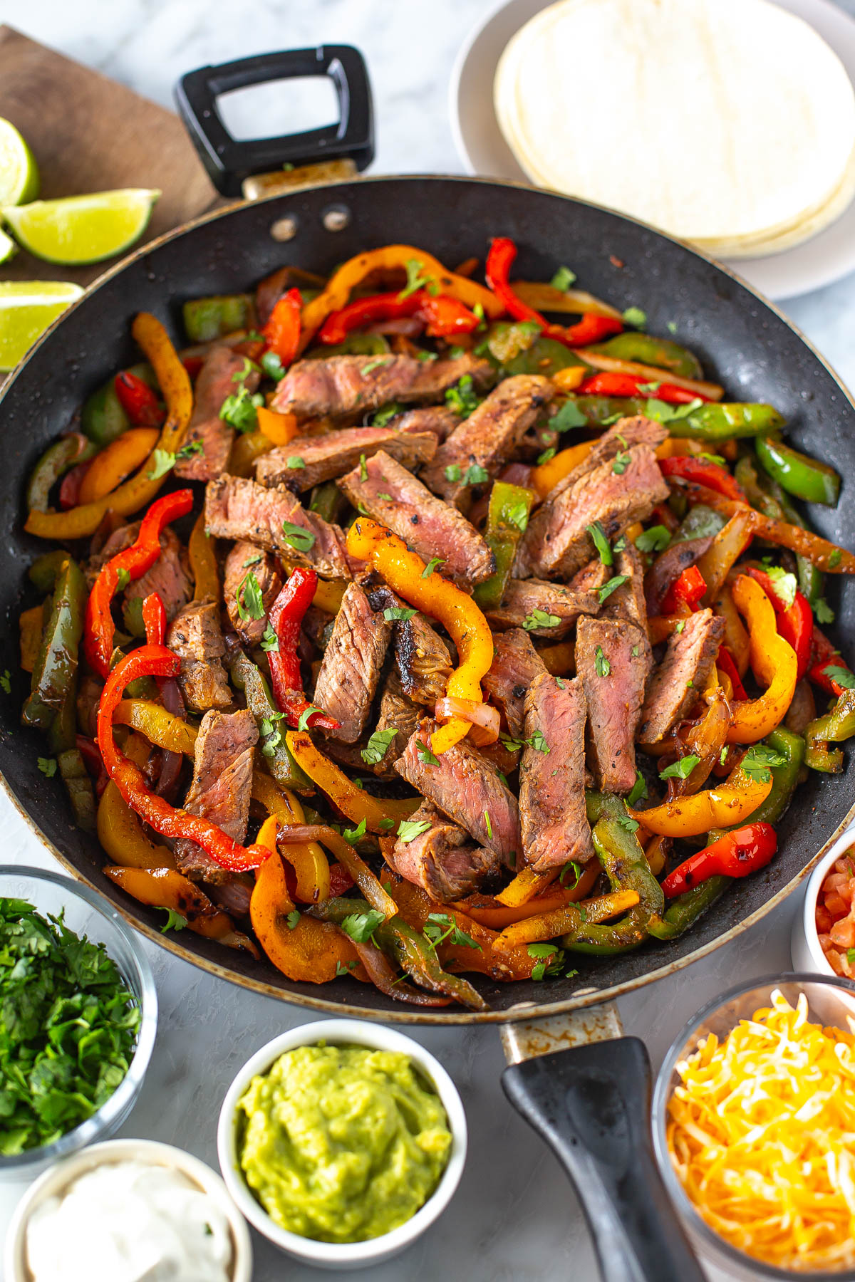 Overhead shot of steak fajitas in a skillet with sliced steak, red and yellow bell peppers, onions, and toppings like guacamole, sour cream, cheese, and salsa