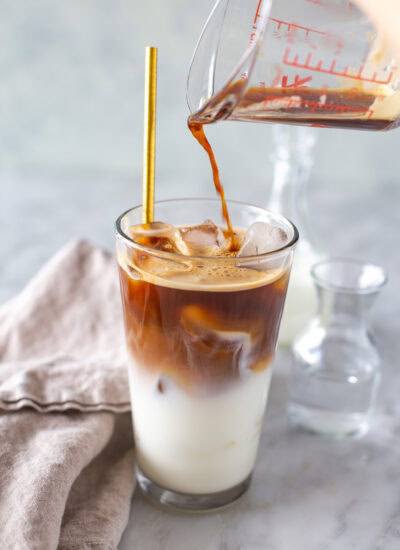 Espresso being poured from a clear measuring cup into a glass of milk and ice, with a gold straw.