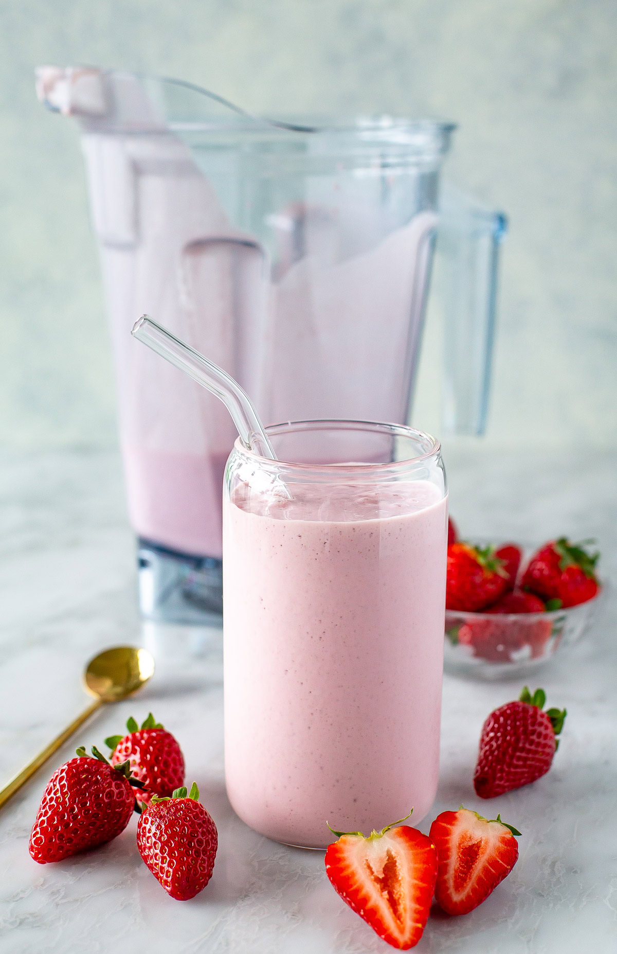 side shot of high protein strawberry smooth in a clear glass with glass straw and a blender pitcher with some strawberries around them