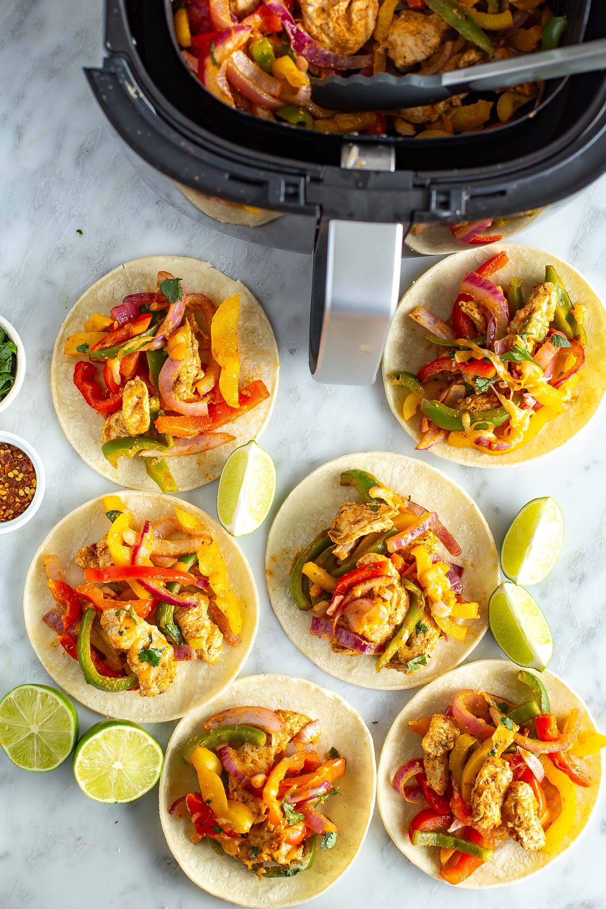 Overhead shot of an air fryer with cooked chicken fajita mixture, surrounded by tortillas filled with chicken, peppers, and onions, garnished with lime wedges