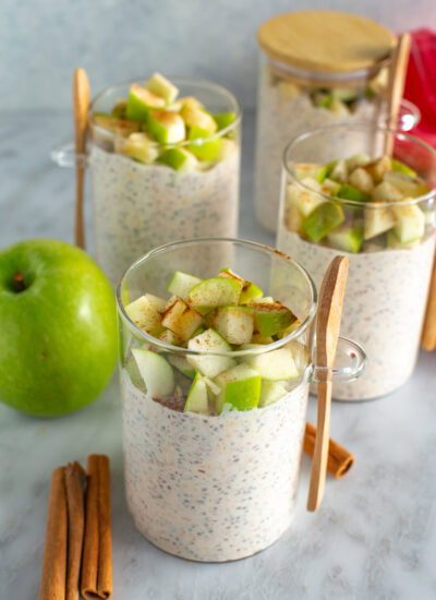 Side shot of four jars of finished Apple Cinnamon Overnight Oats on a counter with a green apple and three cinnamon sticks.
