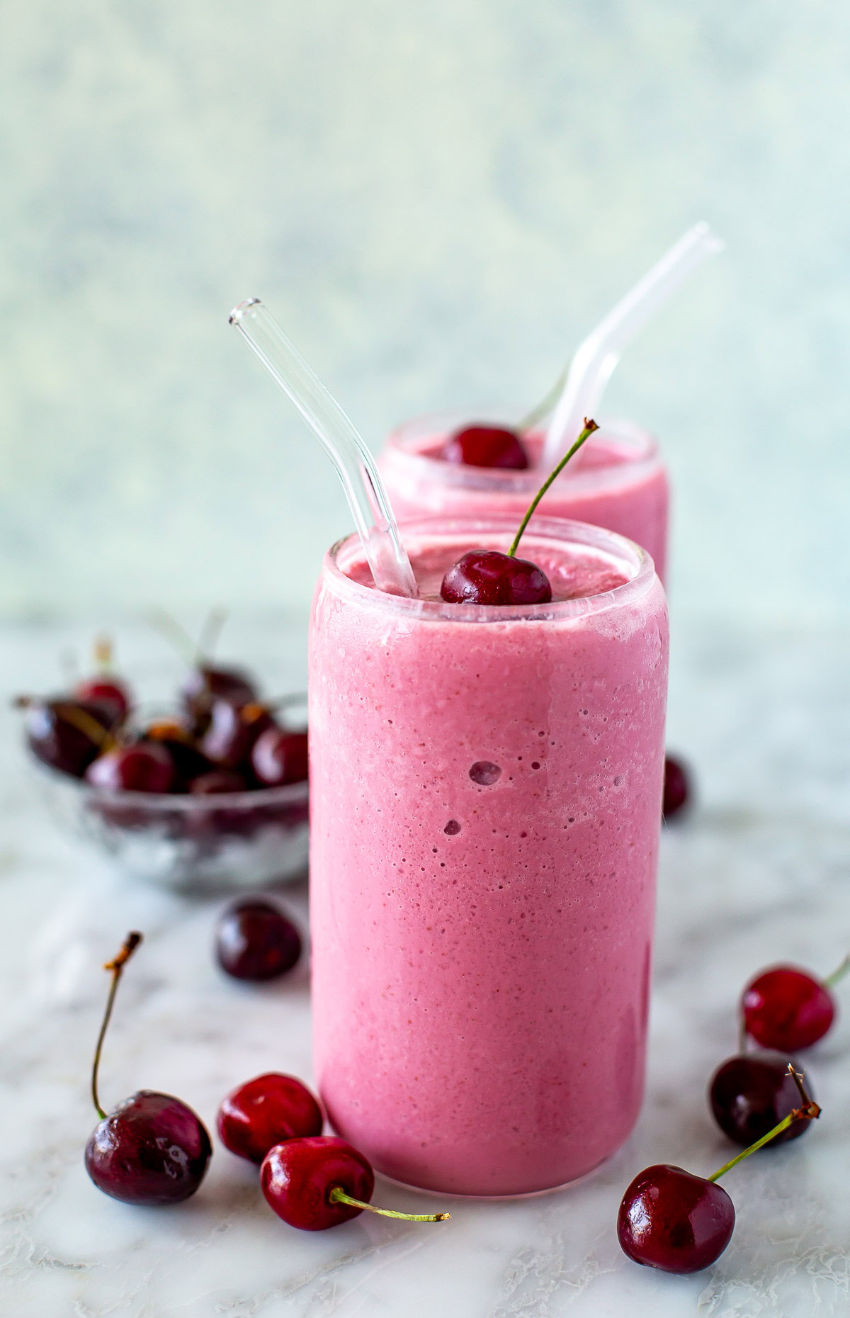 Side shot of two cherry smoothies with straws and fresh cherries scattered around.
