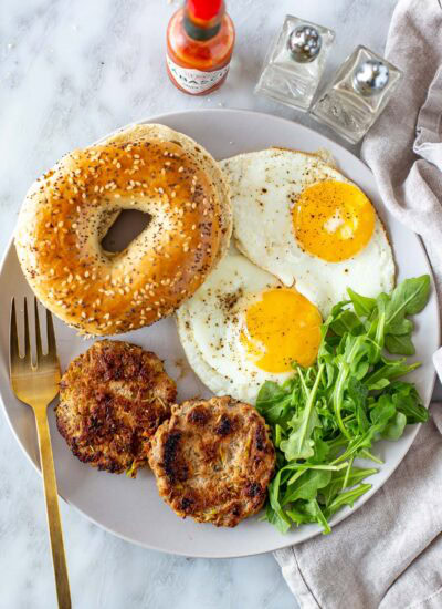 Overhead shot of chicken apple sausage patties, fried eggs, a bagel, and a gold fork.