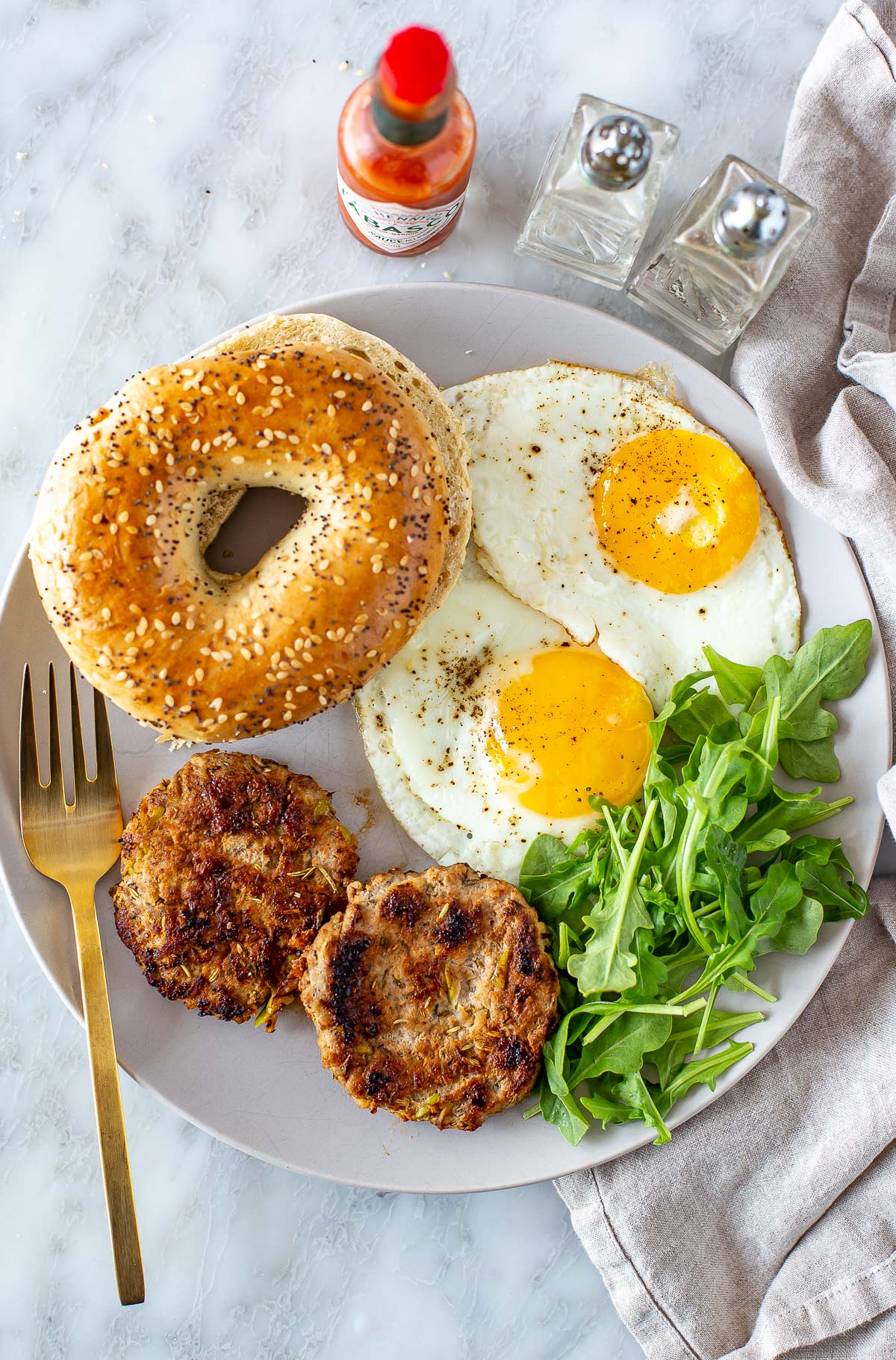 Overhead shot of chicken apple sausage patties, fried eggs, a bagel, and a gold fork.