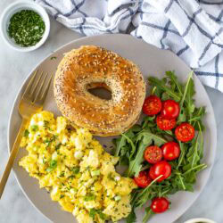Overhead shot of a plate of cottage cheese eggs, a bagel, and an arugula and tomato salad.