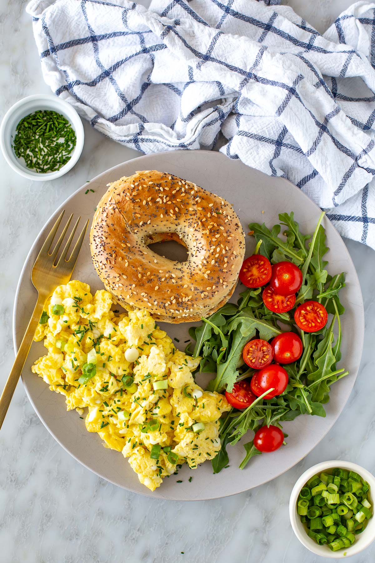 Overhead shot of a plate of cottage cheese eggs, a bagel, and an arugula and tomato salad.