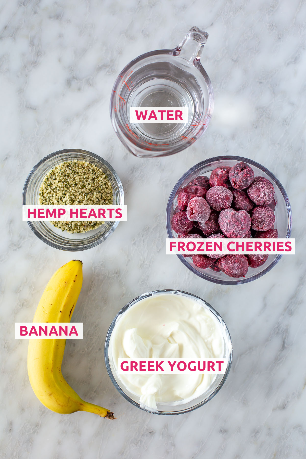 Overhead shot of Easy Cherry Smoothie ingredients, including water, hemp hearts, frozen cherries, banana, and Greek yogurt with labels.