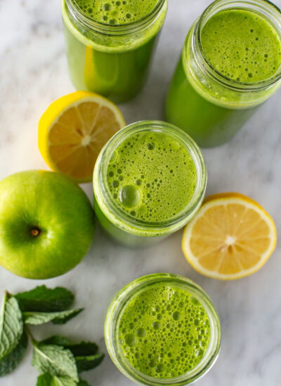 Overhead shot of four glasses of green juice surrounded by lemon and mint.