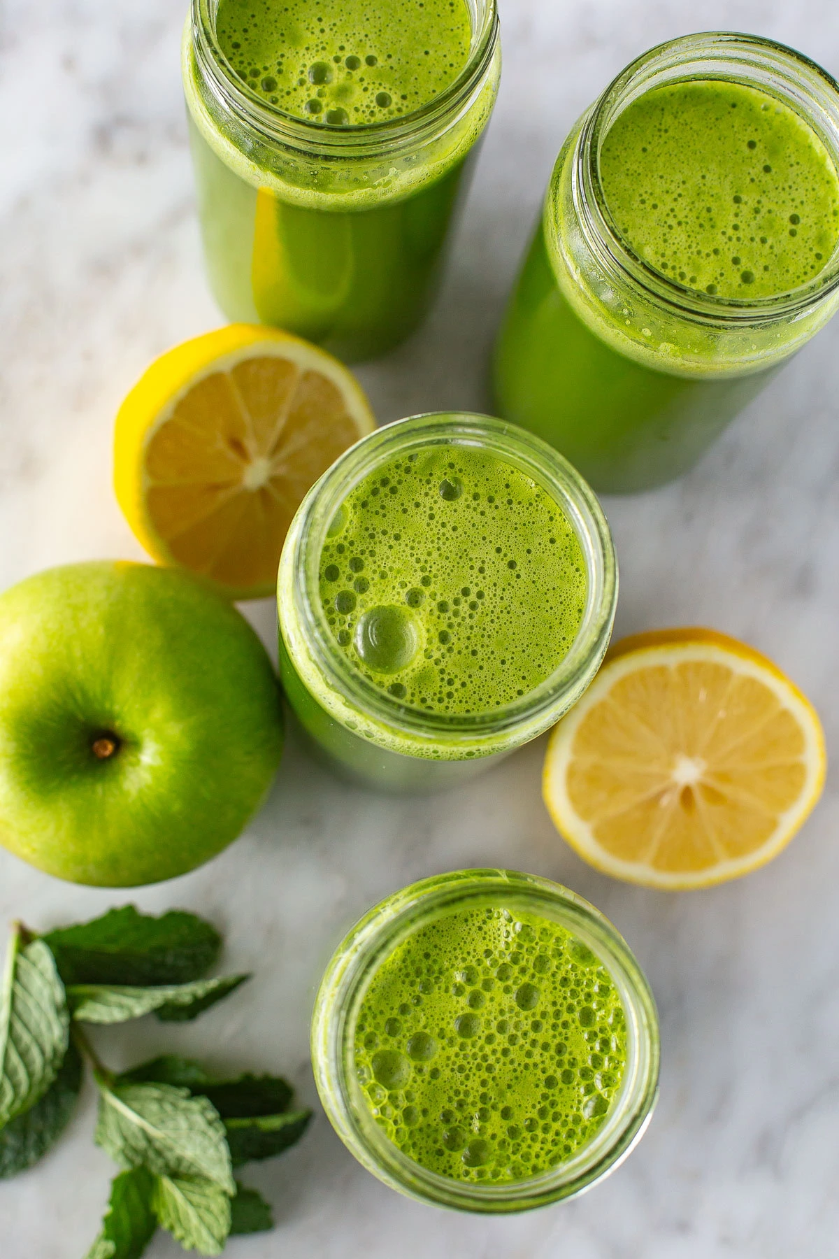 Overhead shot of four glasses of green juice surrounded by lemon and mint.