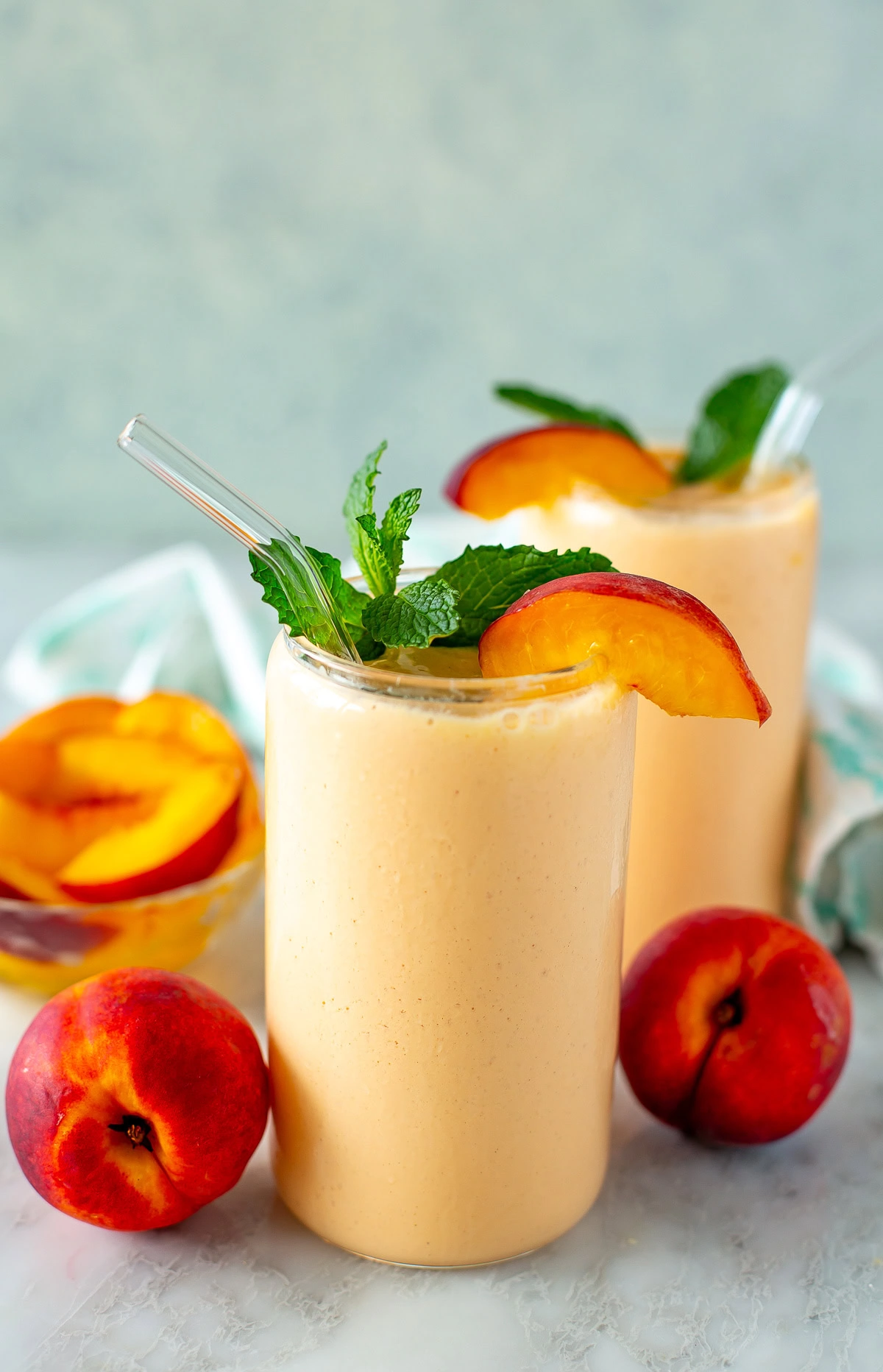 Side shot of a peach smoothie with a straw garnished with peach slices and mint leaves.