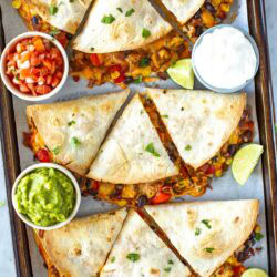 Overhead shot of a sheet pan full of finished quesadillas with side bowls of guacamole and salsa.
