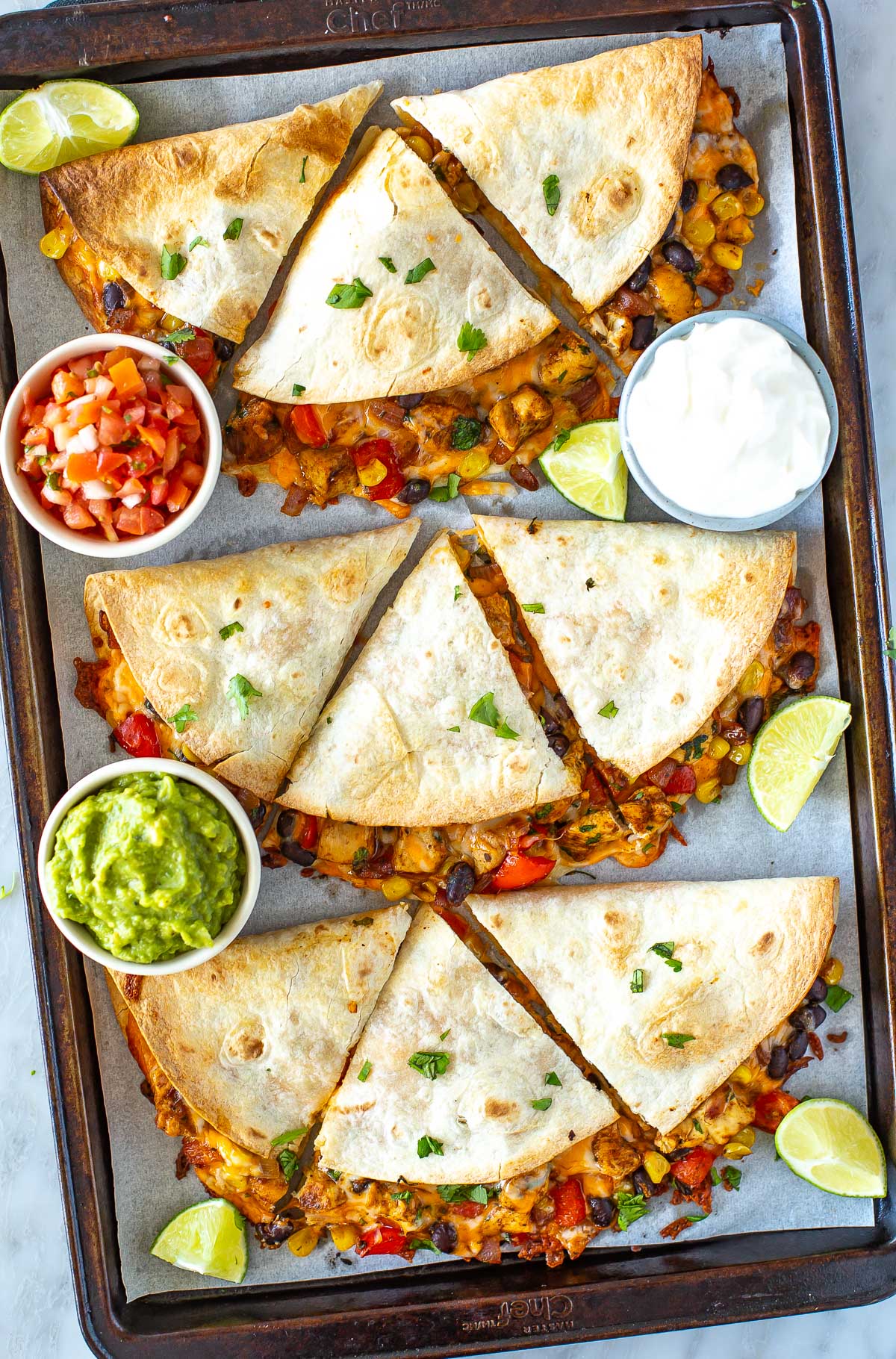 Overhead shot of a sheet pan full of finished quesadillas with side bowls of guacamole and salsa.