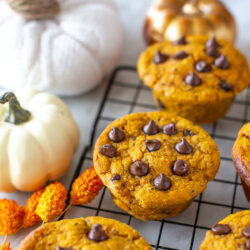 Side shot of several pumpkin protein muffins with chocolate chips on a cooling rack with decorative pumpkins.