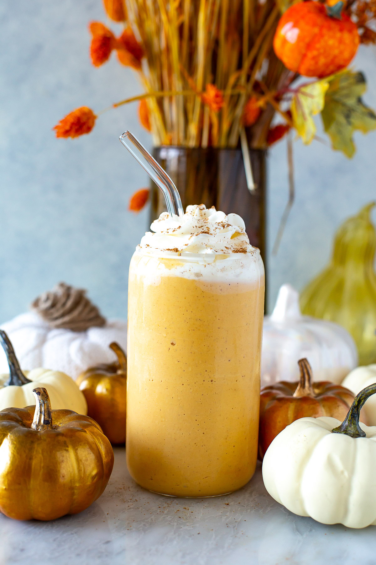 Side shot of a glass of pumpkin spice protein shake with whipped cream and a straw, surrounded by decorative pumpkins.