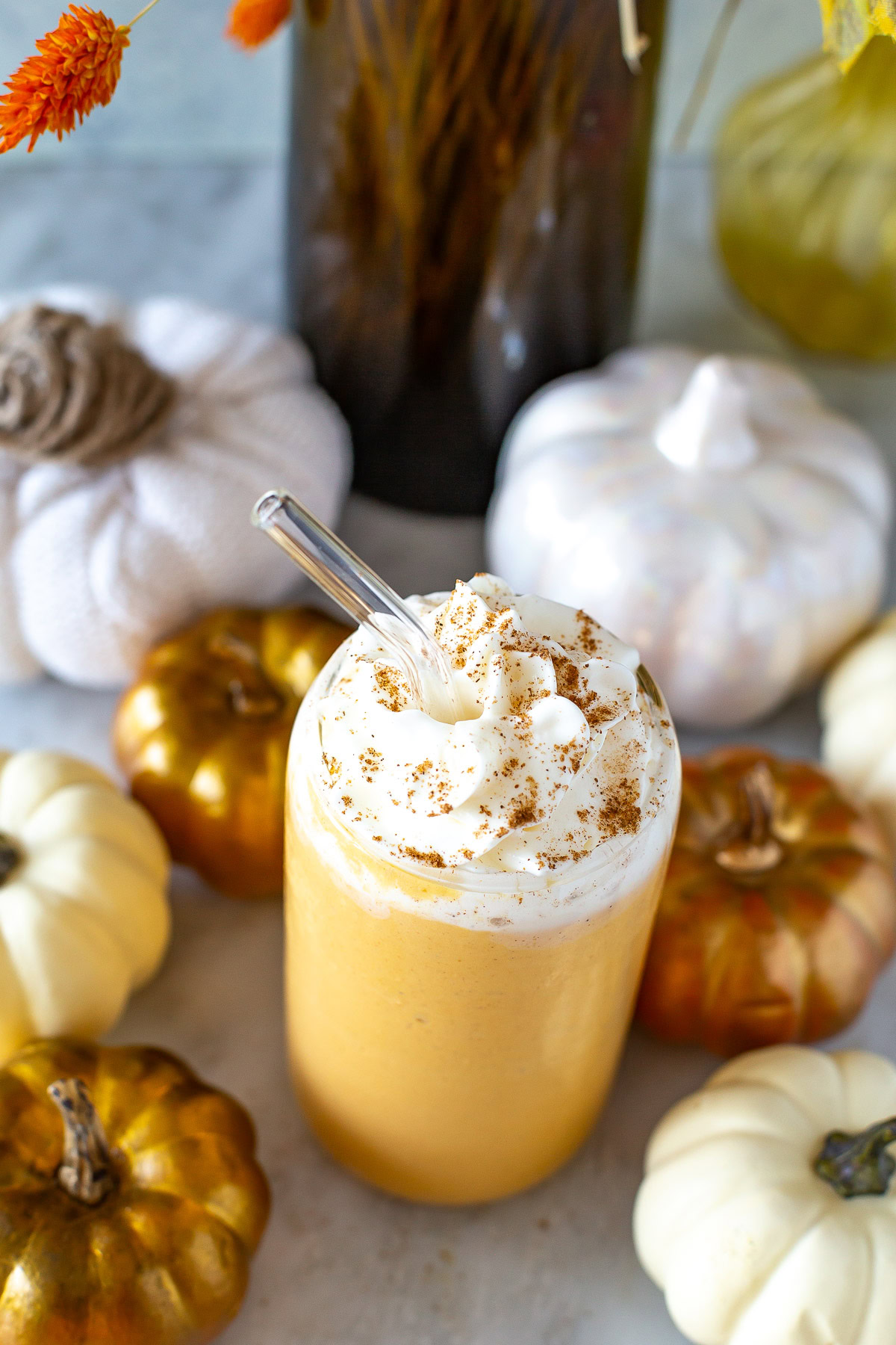 Overhead shot of a glass of pumpkin spice protein shake with whipped cream and a straw, surrounded by decorative pumpkins.