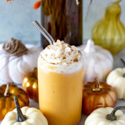 Side shot of a glass of pumpkin spice protein shake with whipped cream and a straw, surrounded by decorative pumpkins.