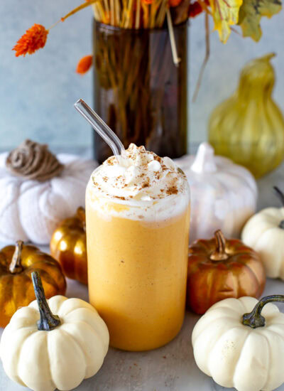 Side shot of a glass of pumpkin spice protein shake with whipped cream and a straw, surrounded by decorative pumpkins.