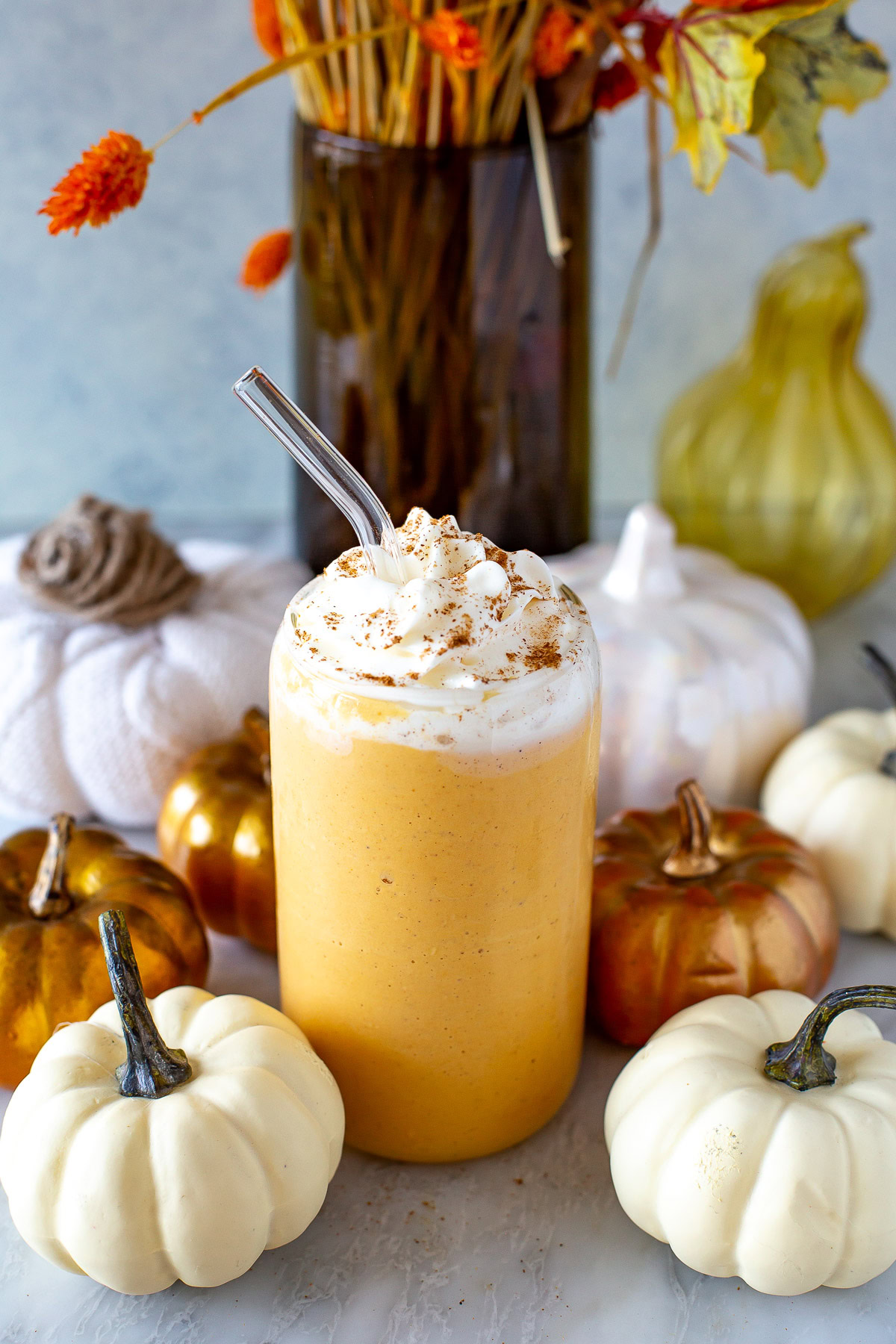 Side shot of a glass of pumpkin spice protein shake with whipped cream and a straw, surrounded by decorative pumpkins.