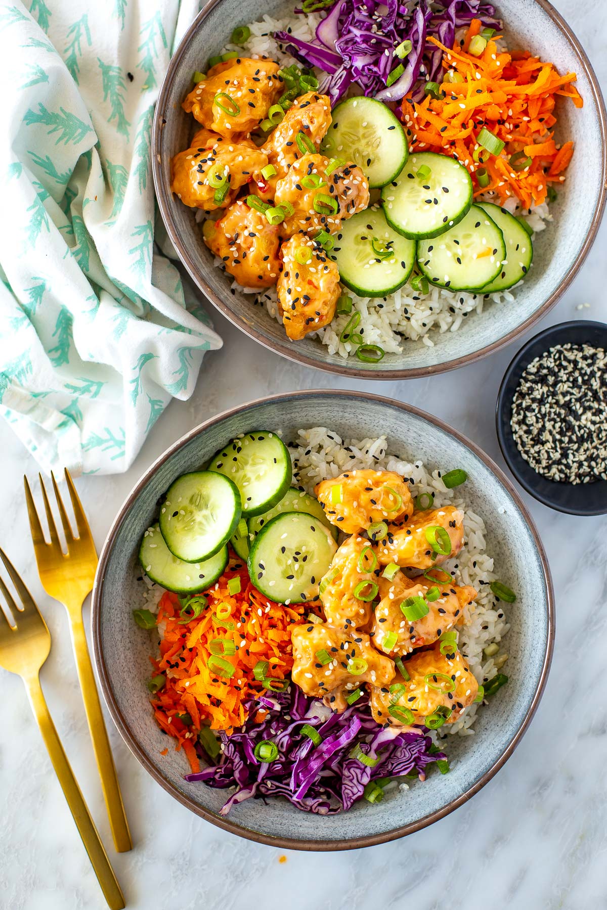 Overhead shot of two bowls of Bang Bang Chicken with a fork and a small bowl of sesame seeds.