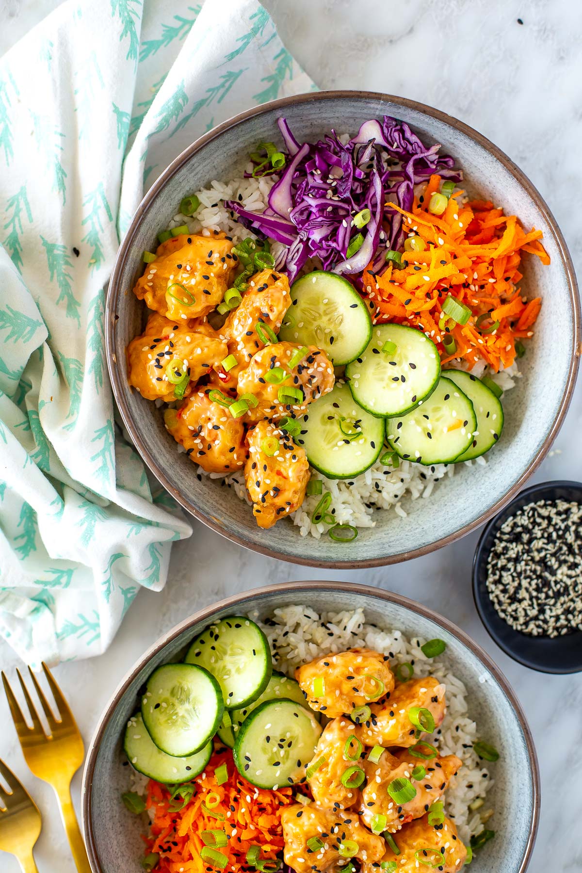 Overhead shot of two bowls of Bang Bang Chicken with a fork and a small bowl of sesame seeds.