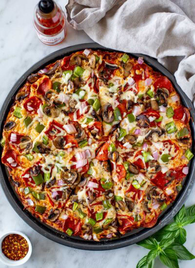 Overhead shot of a whole High Protein Pizza with pepperoni and vegetable toppings in a round pan, sliced into wedges.