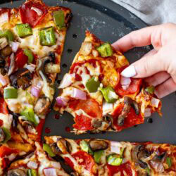 Overhead shot of a hand lifting a slice of High Protein Pizza with pepperoni and vegetable toppings from a round pizza pan.