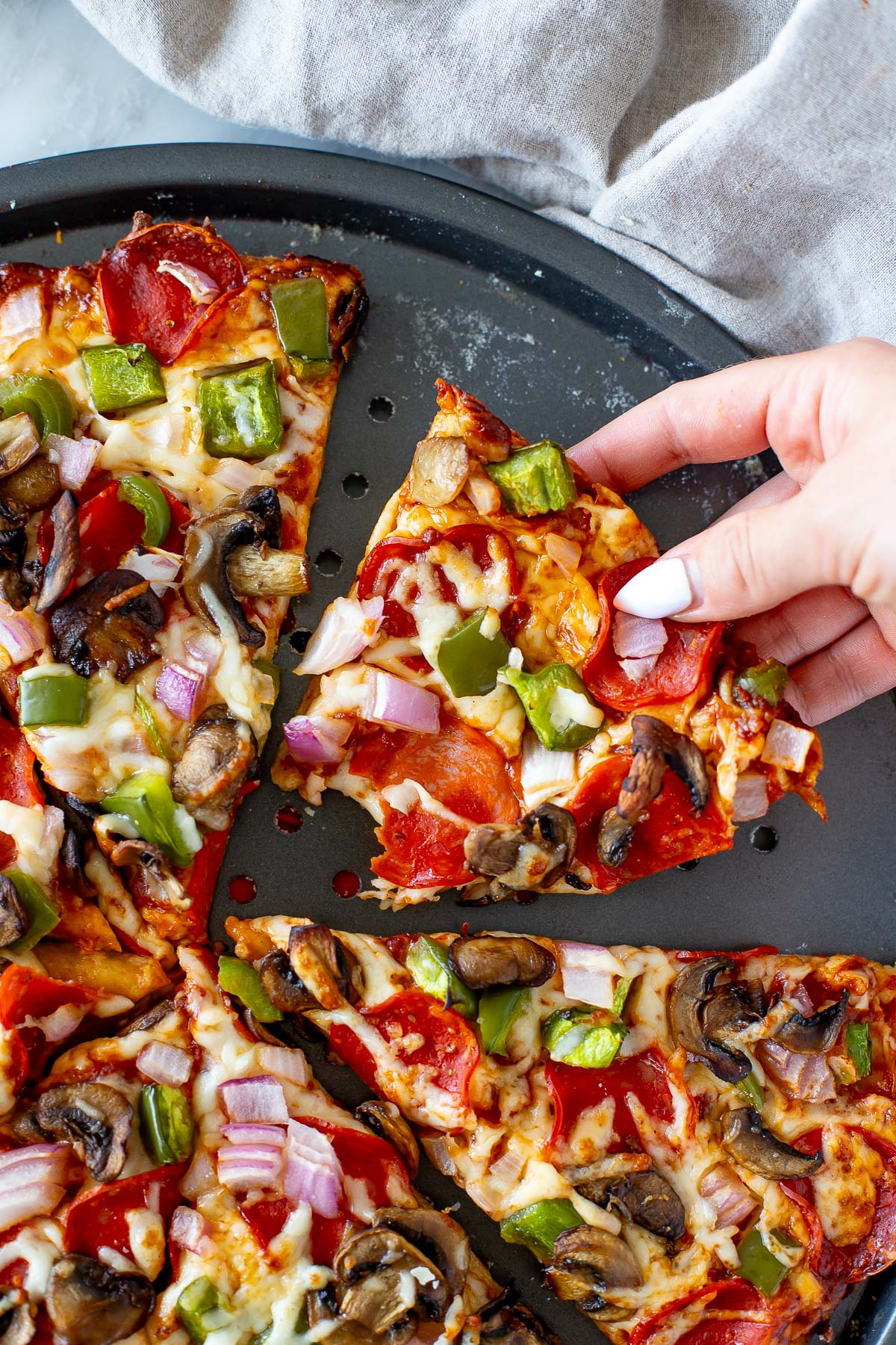 Overhead shot of a hand lifting a slice of High Protein Pizza with pepperoni and vegetable toppings from a round pizza pan.