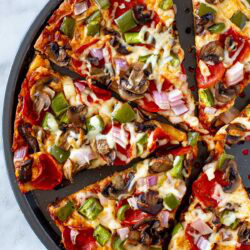 Overhead shot of a sliced High Protein Pizza with pepperoni and vegetable toppings in a round pizza pan.