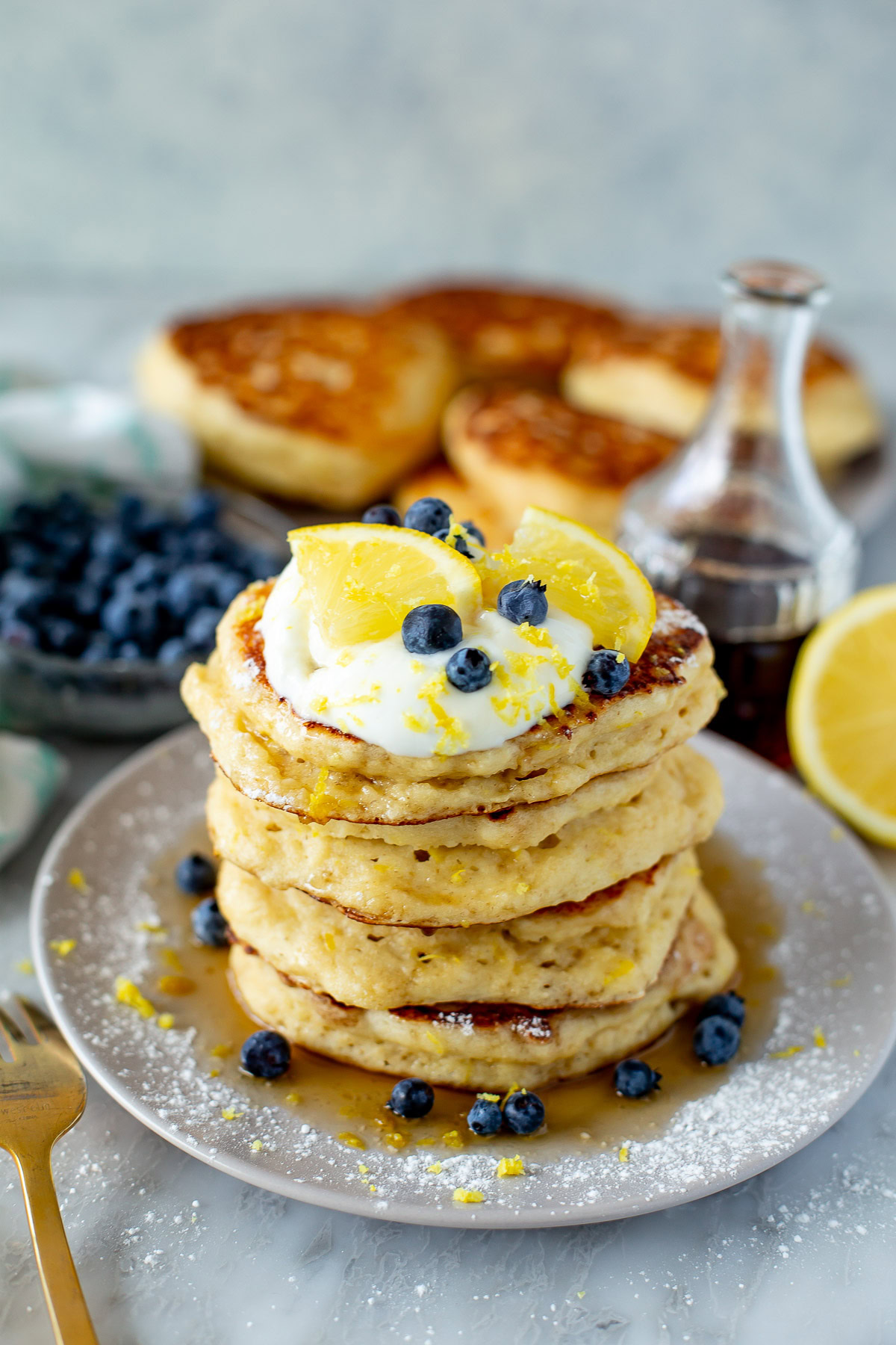 Side shot of a tall stack of Lemon Ricotta Pancakes on a plate, topped with a dollop of cream, blueberries, and a lemon slice.