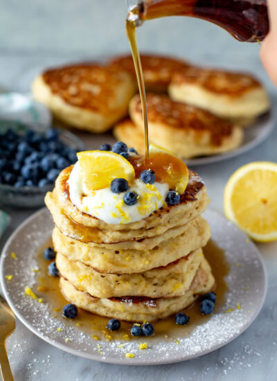 Side shot of a tall stack of Lemon Ricotta Pancakes being drizzled with maple syrup from a glass bottle.
