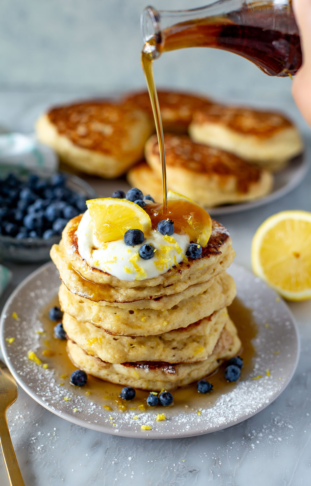 Side shot of a tall stack of Lemon Ricotta Pancakes being drizzled with maple syrup from a glass bottle.