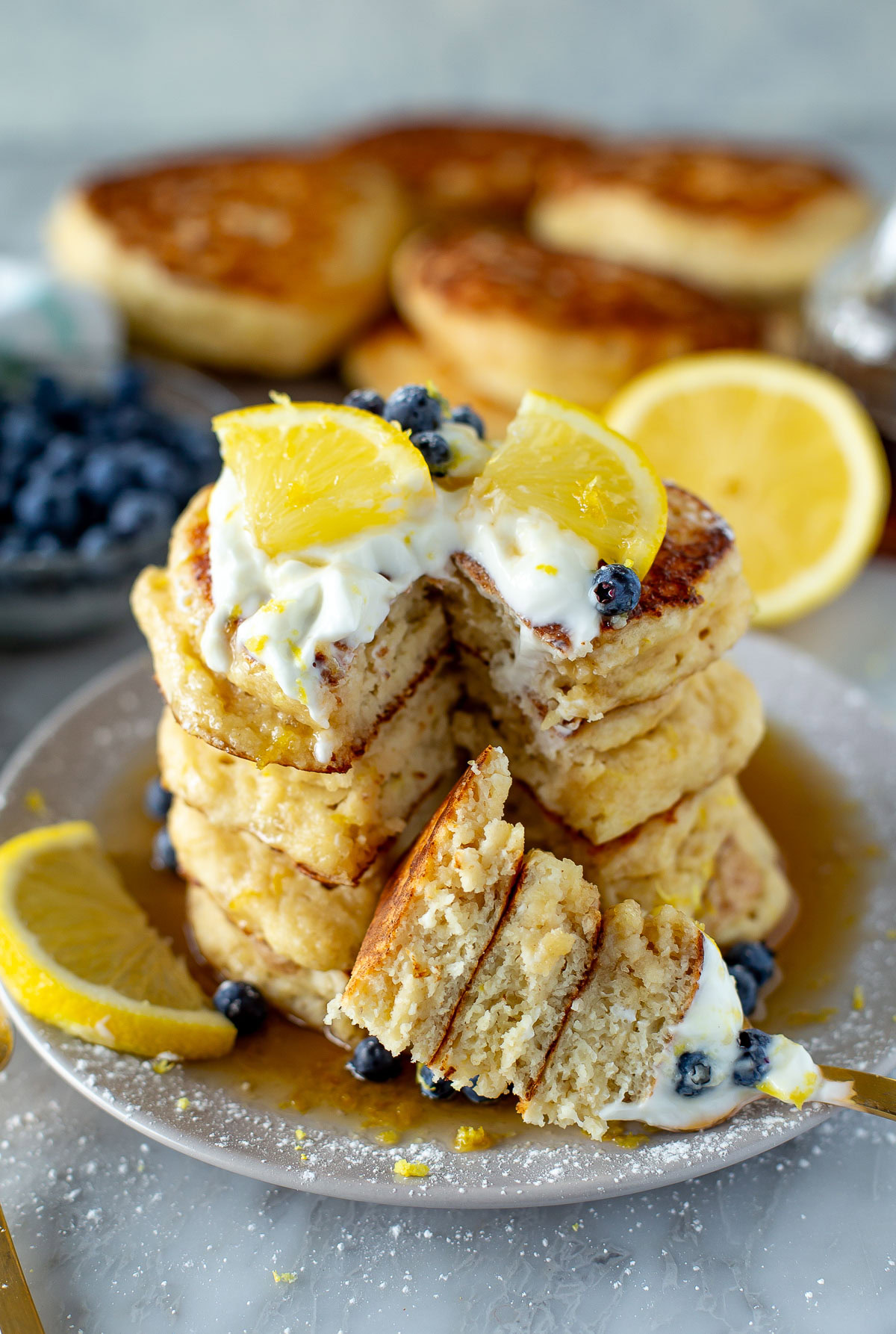 Close-up side shot of a tall stack of Lemon Ricotta Pancakes on a plate, topped with a dollop of cream, blueberries, and a lemon wedge.