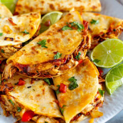 Overhead shot of Cheesy Pulled Chicken Tacos on a white plate with lime wedges and a side of salsa.