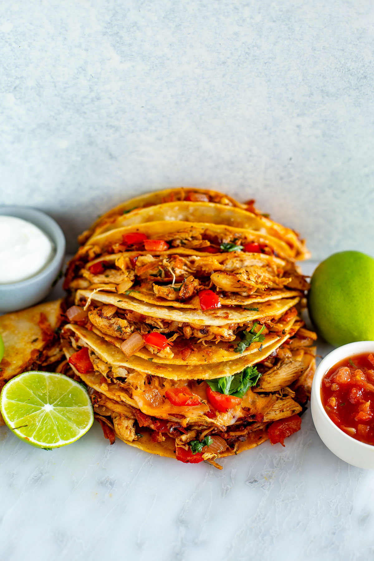 Side shot of a tall stack of Cheesy Pulled Chicken Tacos with sides of sour cream, salsa, and limes.