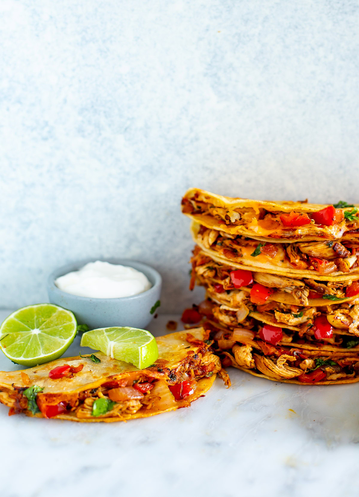 Side shot of a tall stack of Cheesy Pulled Chicken Tacos with a side of sour cream and lime wedges.