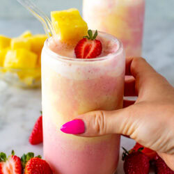 A side shot of a hand holding a glass of a layered strawberry and pineapple smoothie.