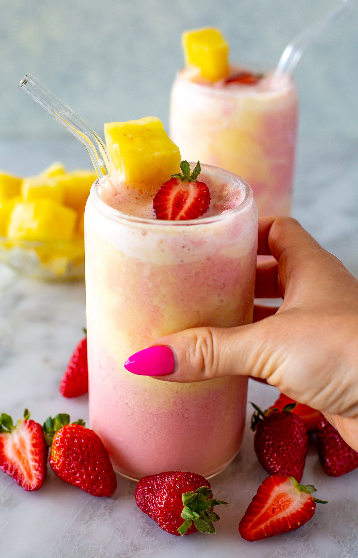 A side shot of a hand holding a glass of a layered strawberry and pineapple smoothie.