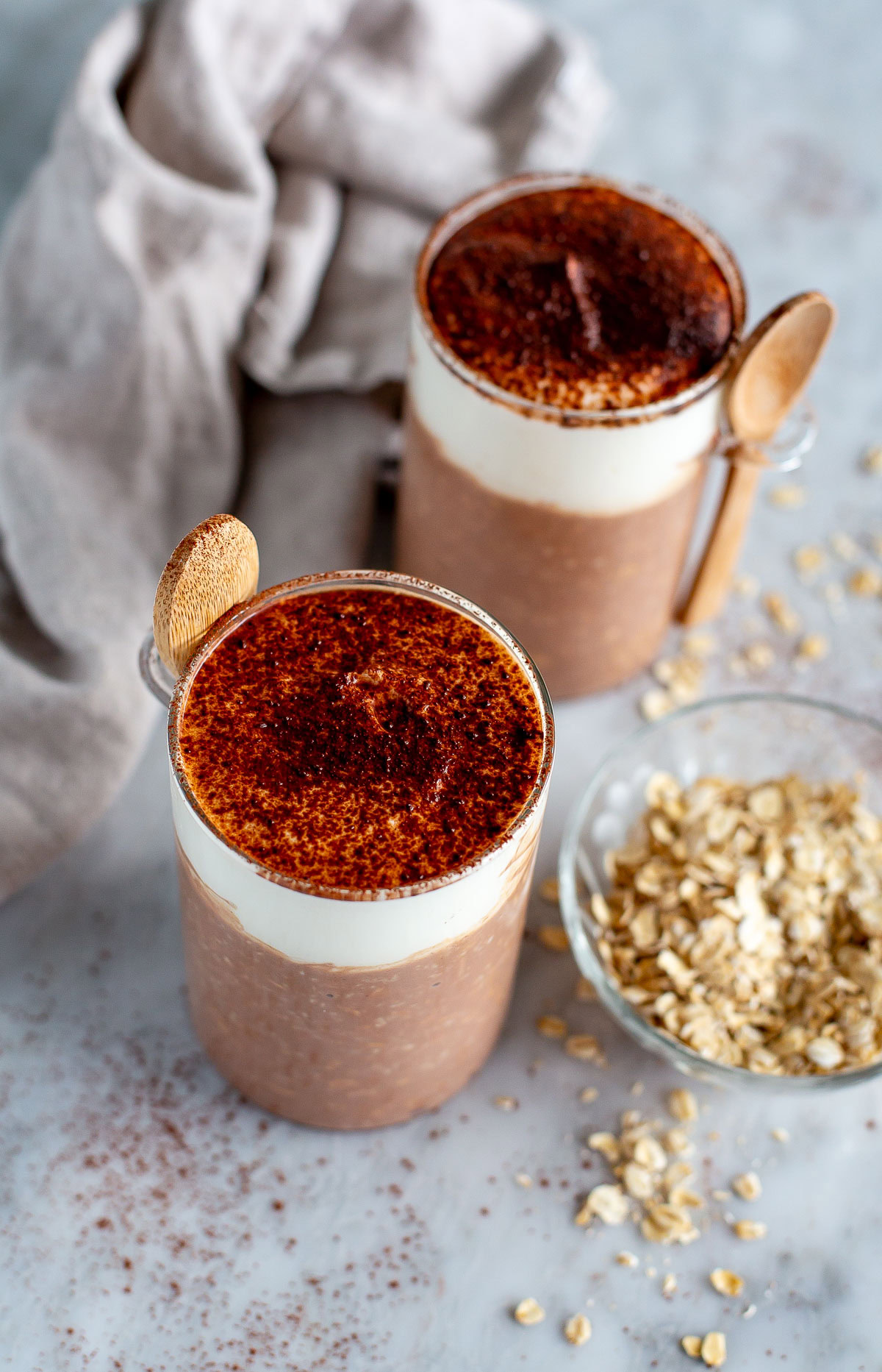 Overhead shot of two jars of Tiramisu Overnight Oats with a cream topping dusted with cocoa powder, next to a small bowl of rolled oats.