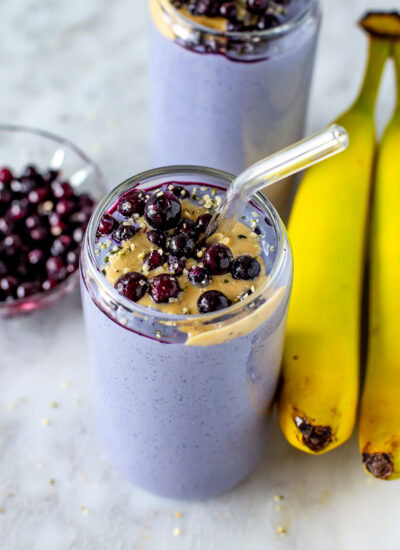 Overhead shot of two purple tofu smoothies topped with blueberries and peanut butter, with bananas on the side.