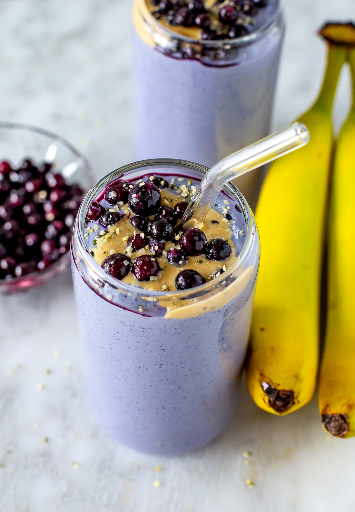 Overhead shot of two purple tofu smoothies topped with blueberries and peanut butter, with bananas on the side.