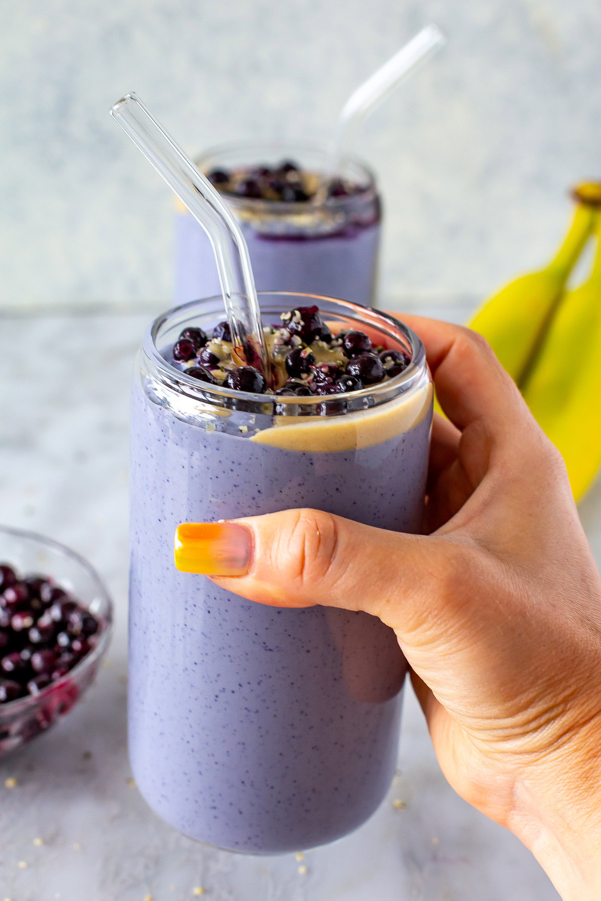 Side shot of a hand holding a purple tofu smoothie topped with blueberries and peanut butter, with two bananas in the background.