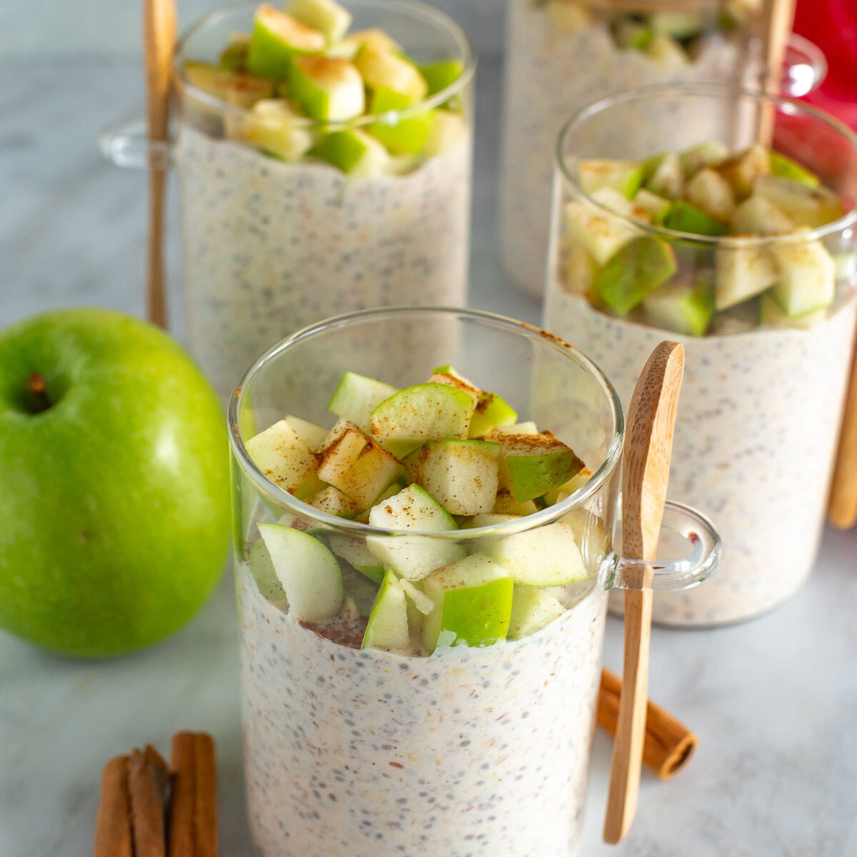 Side shot of four jars of finished Apple Cinnamon Overnight Oats on a counter with a green apple and three cinnamon sticks.