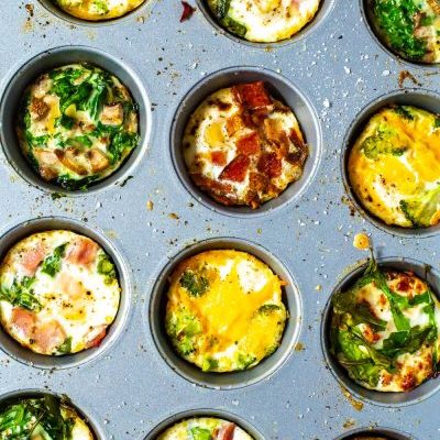 A close-up of a muffin tin filled with 5 different types of baked eggs - mushroom spinach, sundried tomato & arugula, bacon, ham & asparagus, and broccoli cheddar.