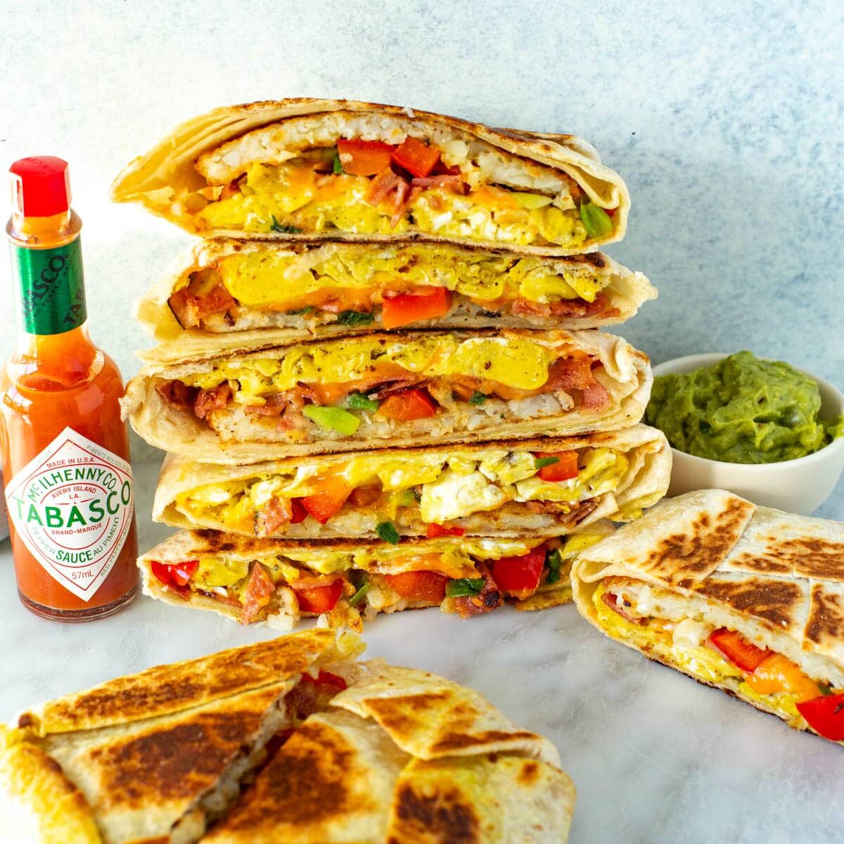 Cut open breakfast crunchwraps stacked together with a side of hot sauce and guacamole.