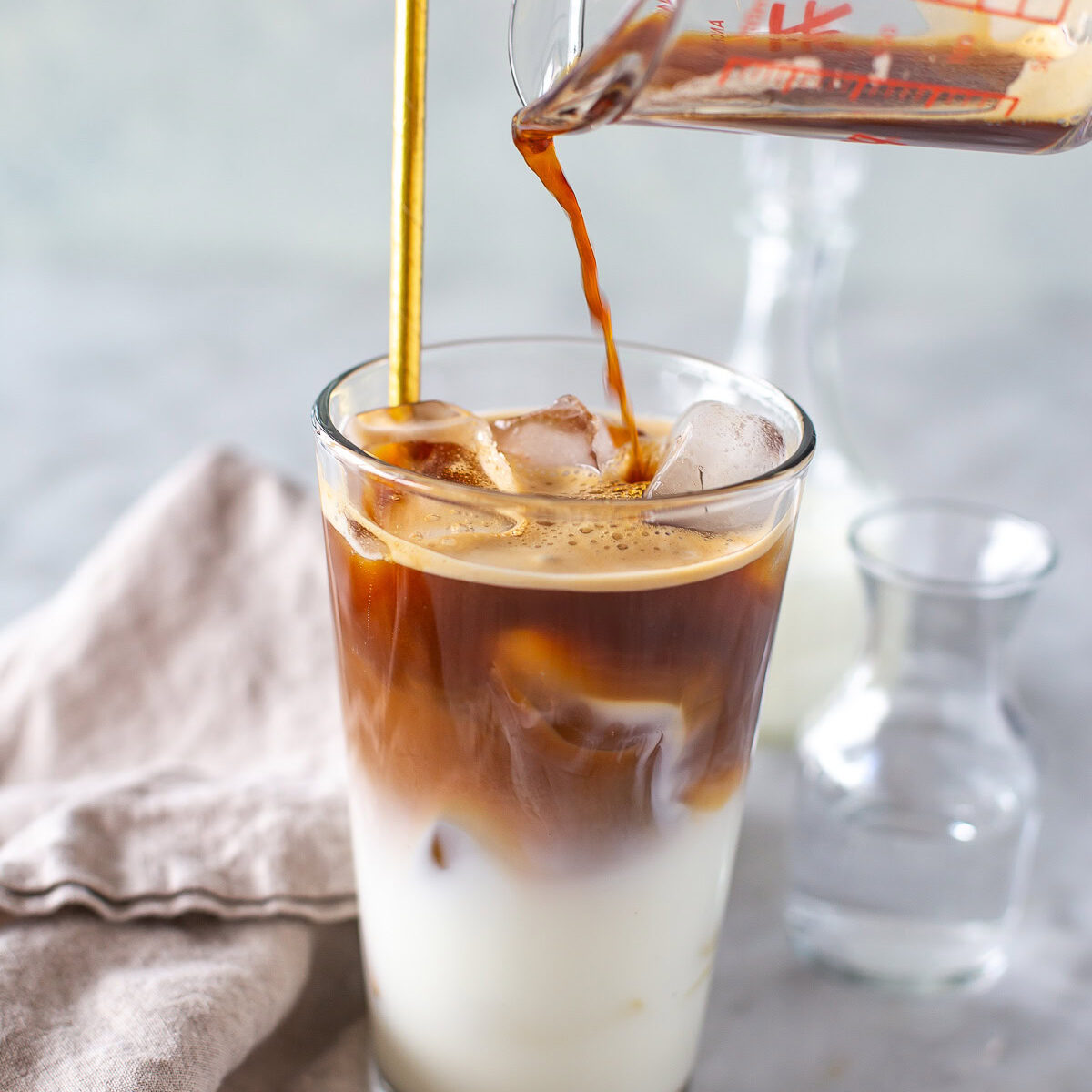 Espresso being poured from a clear measuring cup into a glass of milk and ice, with a gold straw.